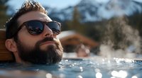 Bearded Caucasian man with sunglasses relaxing in outdoor hot spring pool with steam rising, snowy mountains and cabin in background, wellness retreat atmosphere.