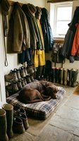 Chocolate Labrador resting on a plaid dog bed in a rustic mudroom with wellington boots, hiking shoes, and hanging outdoor coats.