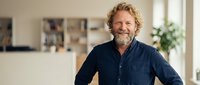 Smiling mature Caucasian man with curly hair and beard in casual navy shirt standing confidently in modern bright office or home workspace interior.