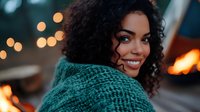 Young African American woman with curly hair smiling warmly in cozy teal sweater with soft bokeh lights creating intimate evening atmosphere for lifestyle content.
