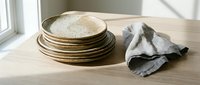 Stack of golden decorative plates with textured surface and crumpled gray linen napkin on wooden table near window, suitable for tableware styling and home decor projects.