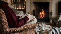 Woman in burgundy sweater reading book by cozy fireplace with warm lighting and comfortable armchair creating peaceful winter evening atmosphere at home.