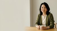 Mature Asian woman with dark wavy hair wearing olive cardigan over white shirt, sitting at wooden table with hands clasped, neutral beige background, professional portrait for business, corporate, or lifestyle use.