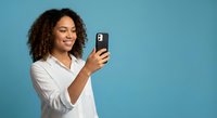 Happy young African American woman with curly hair taking selfie with smartphone on blue background for social media and communication content.
