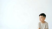 Young Asian male child wearing casual beige t-shirt, smiling while looking to the side against white background. Suitable for educational materials, parenting content, and children's product marketing.