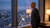 Senior businessman with gray hair and glasses wearing dark suit standing by floor-to-ceiling window overlooking illuminated cityscape at dusk, contemplating urban skyline from high-rise office.