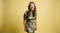 Young Caucasian woman laughing joyfully in tropical print dress against warm yellow background. Studio portrait for lifestyle and fashion content.