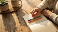 Woman in beige sweater writing in notebook at wooden table with coffee mug and plant in warm natural sunlight for journaling and planning.