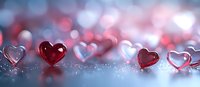 Red and transparent glass hearts scattered on reflective surface with soft bokeh lights in pink and blue tones creating romantic Valentine's Day atmosphere.