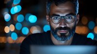 Middle-aged bearded man in glasses illuminated by screen light at night with blue and orange bokeh background, working late on computer, focused expression.