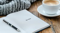 Cozy workspace with open notebook, coffee cup and warm knitted scarf on wooden table. Morning planning and goal setting atmosphere for productive day.
