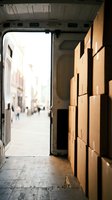 Open delivery van doors revealing stacked cardboard boxes inside with urban street and pedestrians visible in background, suitable for logistics, shipping, e-commerce and distribution concepts.