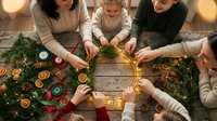 Family making Christmas wreath together at wooden table with dried oranges, pine branches, ribbons, and fairy lights, top view holiday crafting scene.