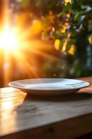 Empty white plate on wooden table with golden sunlight streaming through window, creating warm atmospheric mood with bokeh effect and lens flare.