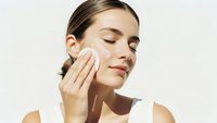Young woman applying facial cream with cotton pad for daily skincare routine and moisturizing treatment on clean background.