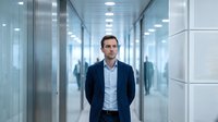 Professional Caucasian businessman in navy suit standing confidently in modern glass office corridor with blurred colleagues in background.