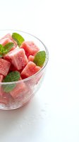 Chilled watermelon cubes with fresh mint leaves in a glass bowl on white surface, perfect for summer beverages, desserts, and food styling.