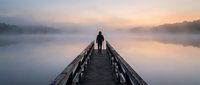 Silhouette of person standing on wooden dock over misty lake at dawn. Peaceful morning scene with fog and soft pastel sky colors for meditation concepts.