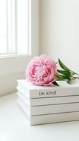 Pink peony resting on a stack of white books with be kind text, soft natural light, minimalist home decor styling.