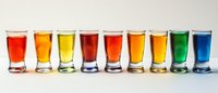 Colorful array of shot glasses filled with vibrant liquids arranged in rainbow sequence on white background, creating striking visual composition for beverage and bar concepts.