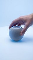Man's hand touching a compact round smart speaker with glowing blue light on white surface, showcasing modern smart home technology.