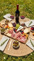 Overhead view of outdoor picnic spread with red wine bottle, cured meats, cheese, fresh grapes, olives, and bread on woven mat over grass for entertaining.