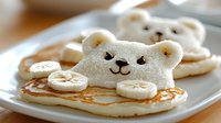 Fluffy bear-shaped pancake with banana slices and chocolate chip details on white plate, creative breakfast for kids featuring cute animal food art presentation.