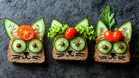 Playful open-faced sandwiches with cucumber eyes and cherry tomato decorations on dark rye bread, arranged on black slate background. Creative food art for kids lunch.