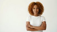 Young African American woman with curly hair in white t-shirt standing confidently with crossed arms against neutral background for business content.
