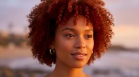 Young African American woman with curly hair smiling at golden hour sunset by ocean. Natural beauty portrait with warm lighting for lifestyle content.