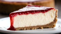 Slice of creamy vanilla cheesecake with bright red raspberry sauce topping on graham cracker crust, served on white plate. Close up view shows smooth texture.