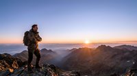 Male hiker with backpack standing on mountain peak at sunrise, overlooking vast valley landscape and distant mountains during golden hour adventure.