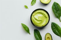 Green avocado cream in black bowl with fresh spinach leaves and sliced avocado on light background, top view. Natural organic skincare and food concept.
