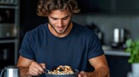 Young Caucasian man with curly hair enjoying healthy breakfast bowl with granola, fruits and yogurt in modern dark kitchen interior, wearing navy t-shirt.