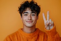 Young Asian man in orange knit sweater showing peace sign gesture against matching orange background, expressing positive cheerful emotion with genuine smile.
