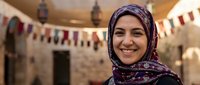 Smiling Middle Eastern woman wearing colorful hijab in traditional courtyard with decorative flags and warm lighting for cultural diversity content.