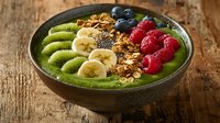 Green smoothie bowl topped with sliced kiwi, banana, raspberries, blueberries and crunchy granola on rustic wooden surface. Fresh healthy breakfast.