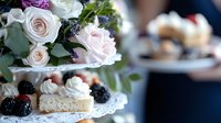 Elegant tiered dessert stand with fresh pink roses and mini pastries topped with whipped cream and blackberries, creating romantic afternoon tea atmosphere.