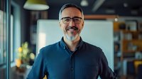 Confident mature businessman with gray beard and glasses wearing navy blue shirt smiling in modern office space. Natural window lighting creates professional atmosphere.