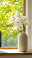 White orchid flowers in modern beige vase on windowsill with bright green summer foliage background through window, creating serene home atmosphere.