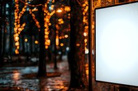 Empty white billboard mockup at night with blurred bokeh lights and trees in background, wet street reflection creates magical evening atmosphere.