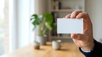 Adult male hand holding blank white business card in modern office workspace with potted plant and desk accessories visible in background.