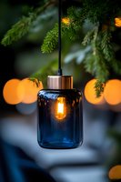 Modern blue glass pendant lamp with glowing Edison bulb hanging under pine branches, orange bokeh lights creating cozy atmospheric mood for home decor.