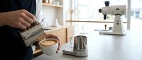 Professional barista pouring steamed milk creating latte art in white cup at modern coffee shop counter with espresso machine and metal pitcher.