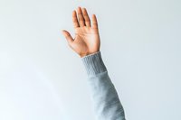 Raised hand in gray sweater sleeve against light background showing greeting, voting, participation or volunteering gesture. Minimalist composition.