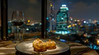 Gourmet dessert profiteroles on dark plate with red wine glass against blurred city nightscape and illuminated skyscraper view through restaurant window.