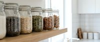 Glass jars with various grains, seeds, and legumes organized on wooden shelf in bright kitchen with white subway tile backsplash for pantry storage.