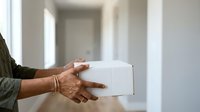 Senior woman with brown skin receiving white cardboard box delivery in modern hallway, wearing olive green sweater with gold bracelet, logistics and parcel service concept.