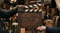 Filmmaker holding vintage film clapperboard on movie set with warm lighting and production equipment in background, used for cinema production and filmmaking projects.