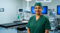 Smiling female surgeon in green scrubs and surgical cap standing confidently in modern operating room with medical equipment and monitors in background.